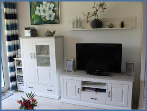 Der Wohnbereich ist modern und besitzt eine komfortable Sitzecke und einem bequemen Sessel. Ein Flachbildfernseher und eine Musikanlage in einer hellen Wohnwand runden das Bild ab.